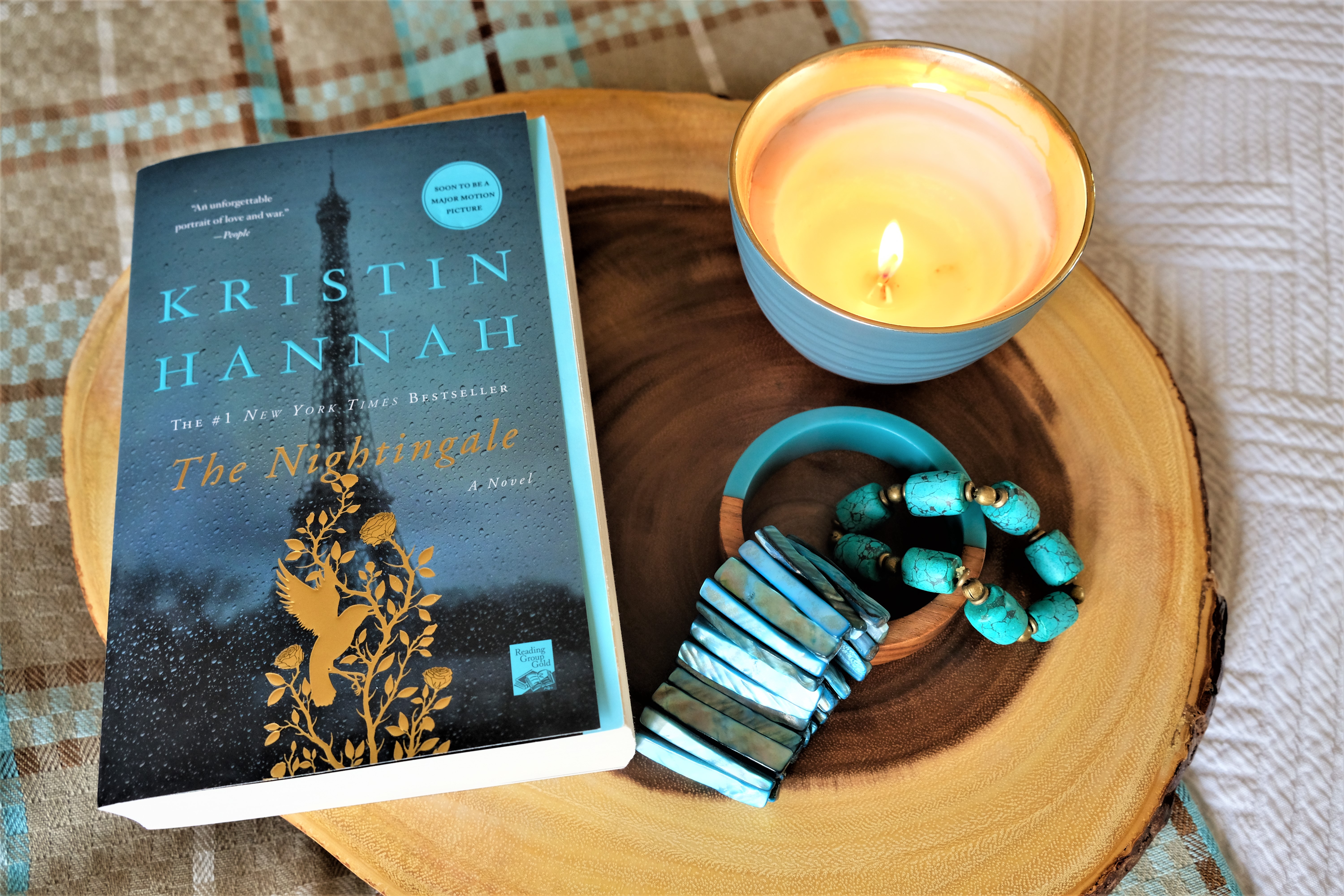 The book is The Nightingale by Kristen Hannah placed on a wooden board with an accessory of a candle and some bracelets